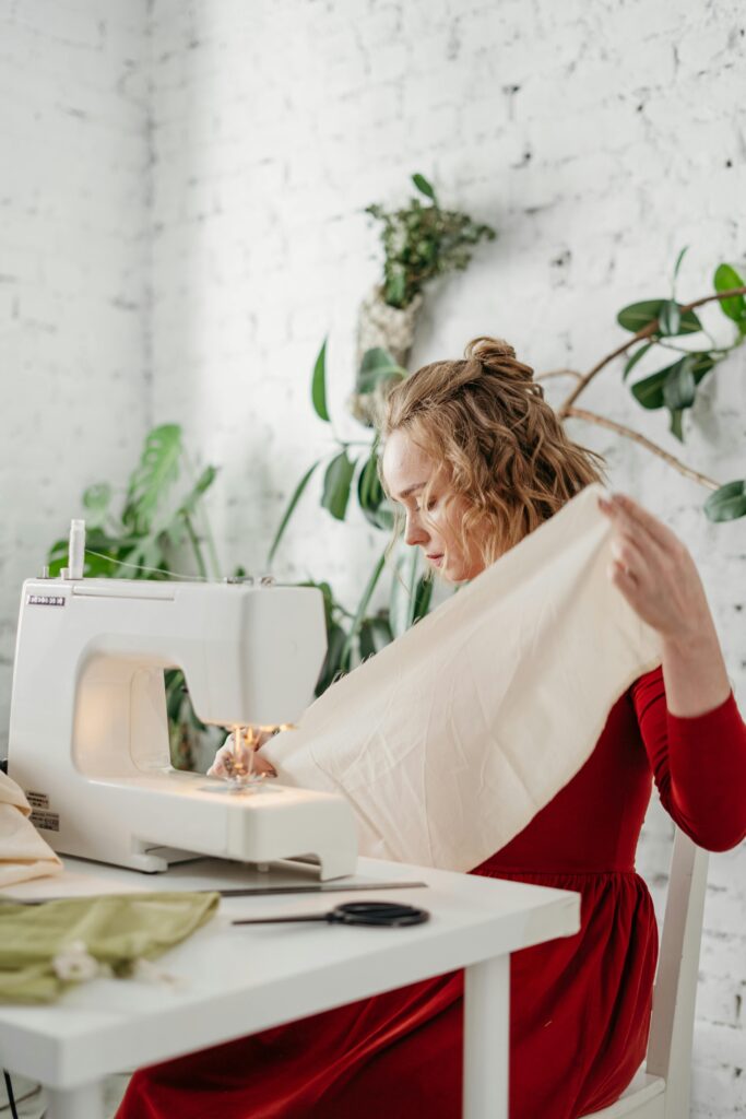 Woman in red dress sewing with a machine in a bright room surrounded by plants.