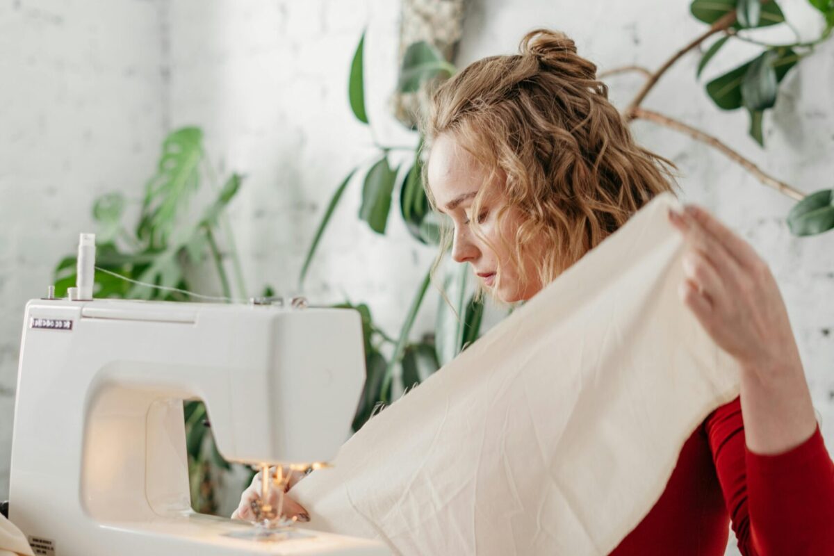 Woman in red dress sewing with a machine in a bright room surrounded by plants.